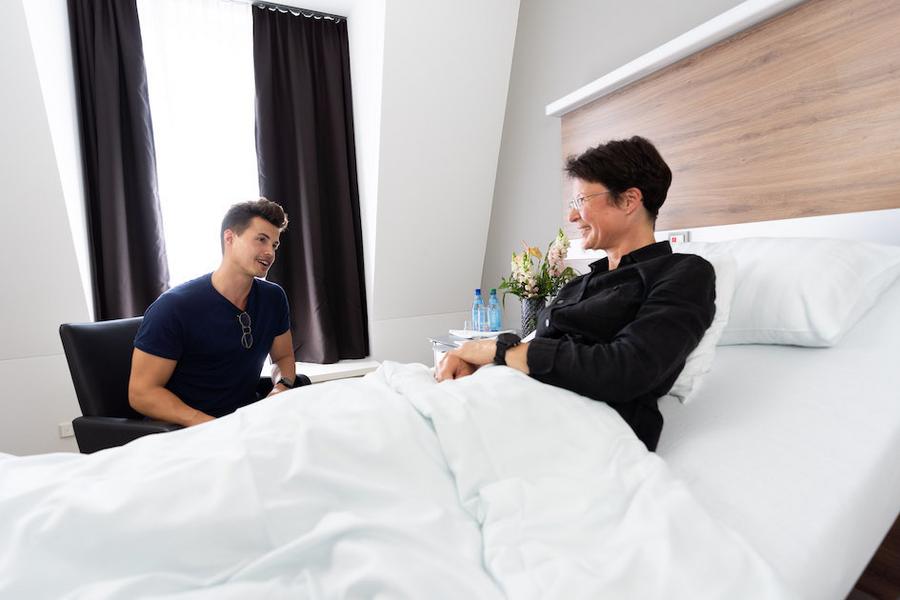 Ältere Patientin in ihrem Krankenbett, ein junger Mann besucht sie, sitzt links von ihr auf einem Stuhl, beide lächeln
