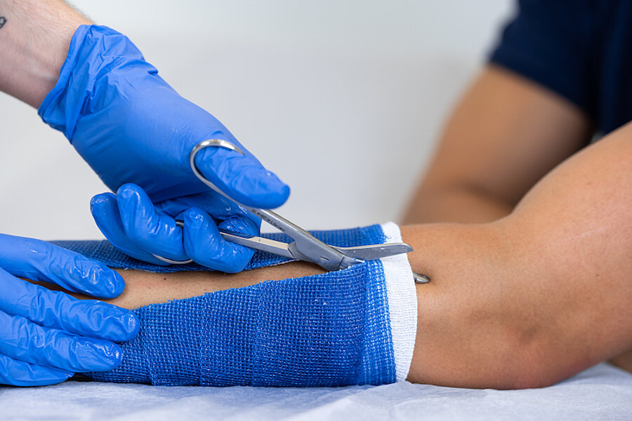 Gipsverband wird vom Unterarm eines Patienten entfernt, behandelnde Hände in Handschuhen, aufgeschnittener Gips und Schere sichtbar