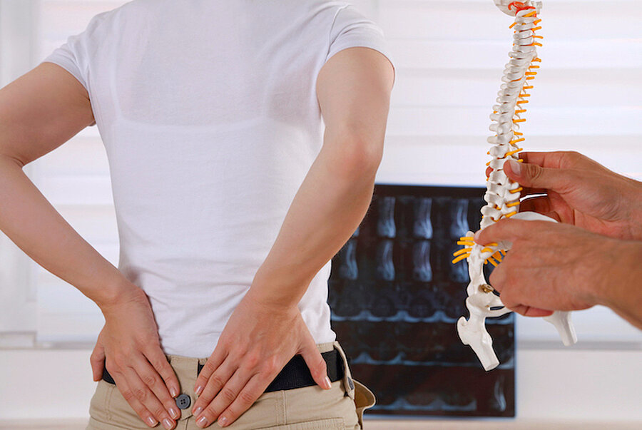 Patientin von hinten, beide Hände am unteren Rücken, Röntgenbild im Hintergrund, vorn rechts im Bild Hände eines Arztes mit Wirbelsäulenmodell