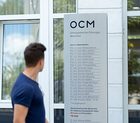 Junger Patient, seitlich zu sehen, blickt auf das Eingangsschild der OCM Orthopädische Chirurgie München