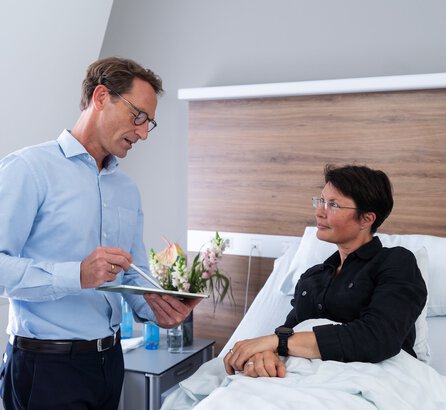 Arzt, links im Bild stehend, spricht mit einer Patientin im Krankenbett, rechts im Bild, beide im mittleren Alter