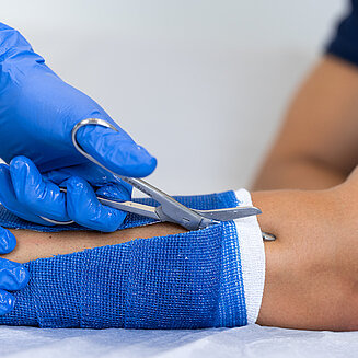 Zwei Hände mit blauen Arzthandschuhen, Schere, Gipsarm eines Patienten wird aufgeschnitten