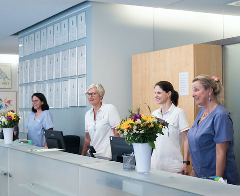 4 Mitarbeiterinnen in weißer und blauer medizinischer Berufskleidung stehen nebeneinander in einer Reihe am Empfang der Orthopädischen Chirurgie München, alle lächeln freundlich