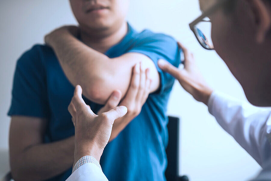 männlicher Patient im T-Shirt, nicht vollständig sichtbar, linker Arm und Ellenbogen in Bildmitte, Arzt untersucht Arm und Schulter