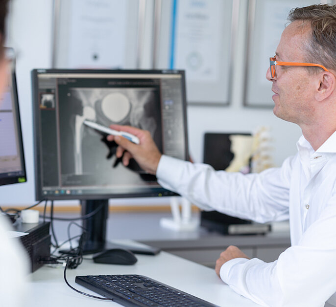 Prof. Robert Hube, hip specialist at the OCM Orthopedic Surgery Munich, talking to a patient, both are seated and looking at a monitor showing an x-ray image of a hip joint, Prof. Hube is pointing at the x-ray image with a pen