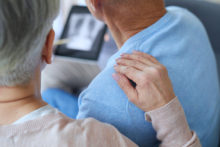 Älteres Paar beim Arzt, Rückansicht, beide blicken auf Röntgenbild, Hand der Frau liegt auf linker Schulter des Mannes